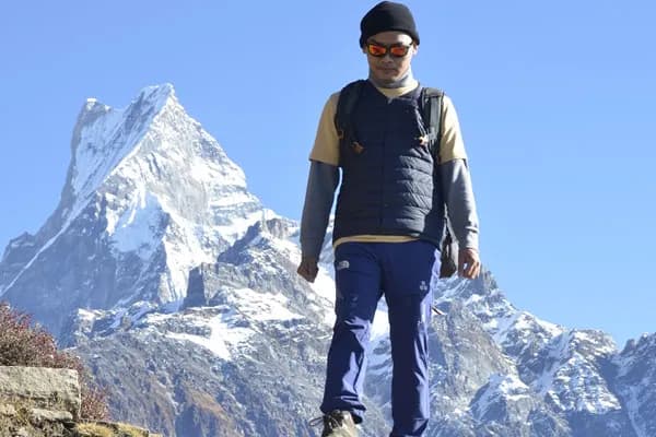 A beginner trekker wearing sunglasses and warm trekking attire walking along a mountain path with Mount Machhapuchhre (Fishtail) towering in the background
