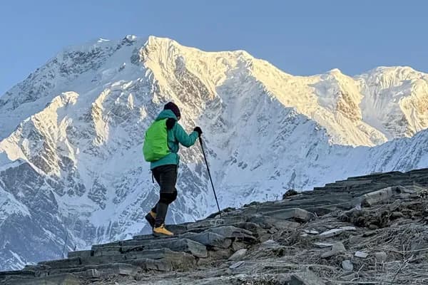 A solo hiker with green backpack and trekking poles climbing stone trail stairs with massive snow-capped mountains illuminated by sunlight ahead