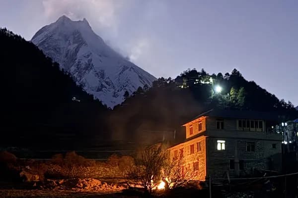 A mountain teahouse illuminated with warm indoor lighting and an outdoor campfire at night under a clear sky with snow-capped Mount Manaslu glowing in the background