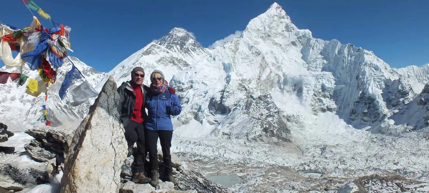 A senior trekking couple wearing warm winter gear and sunglasses posing on a snowy mountain summit against a clear blue sky
