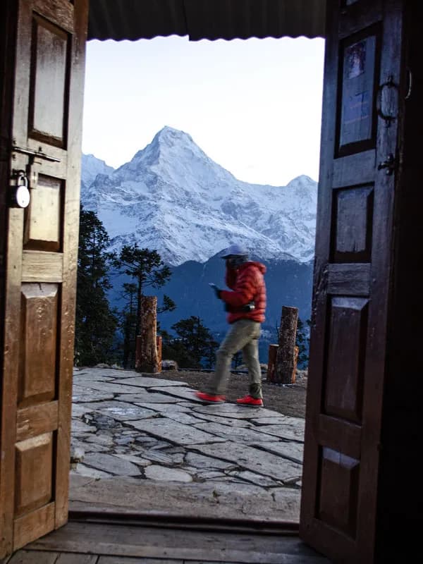 A hiker in a red jacket walking along a stone patio framed by open wooden doors, with a high snow-capped Himalayan peak in the background