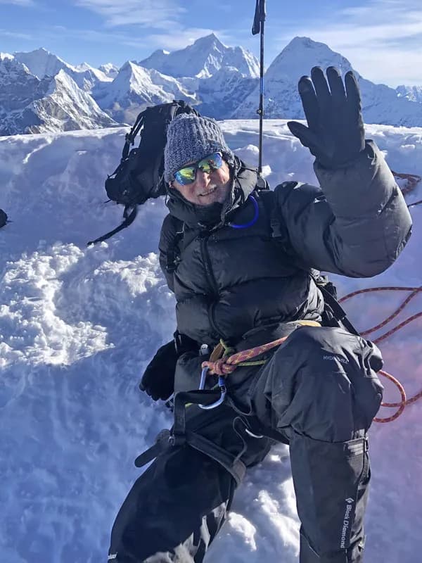 A climber in full cold-weather gear, harness, and ropes waving from a snowy peak summit with distant Himalayan summits behind