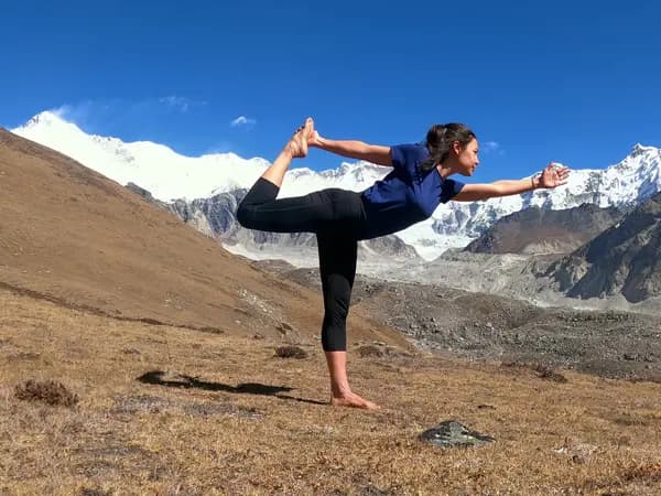 A trekker holding Natarajasana (Dancer Pose) outdoors with snow-covered Himalayan mountains behind her.
