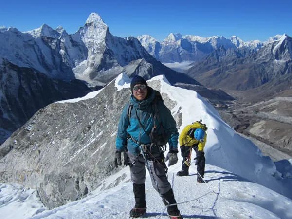 A climber standing on a sharp snow ridge with Ama Dablam's iconic summit rising prominently in the background under a blue sky