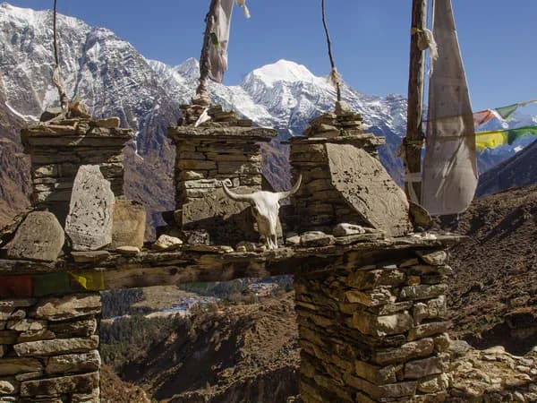 Traditional stone gateway decorated with Mani stones, prayer flags, and an animal skull against snow-capped Himalayan peaks in Tsum Valley