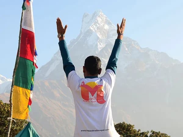 A trekker wearing a Mardi Himal Yoga Trek t-shirt stands with raised arms facing the iconic peak of Mount Machhapuchhre (Fishtail) alongside colorful prayer flags.