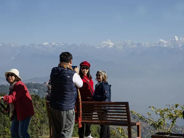 Tourists standing near a wooden bench on a hillside, taking photos of the Himalayan landscape