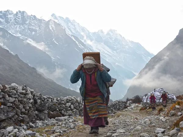 A local Sherpa porter in traditional apron carrying a heavy load using a head strap along a rocky mountain trail in Rolwaling