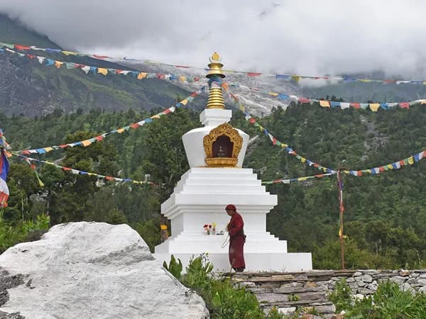 A Buddhist monk praying at a white chorten decorated with prayer flags against a lush mountain backdrop in Tsum Valley