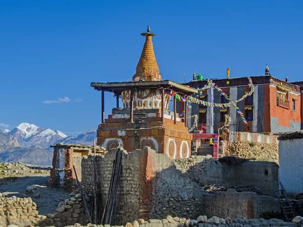 Traditional Buddhist stupa and monastery with colorful prayer flags in Upper Mustang, Nepal, against a clear blue sky and distant snow-capped mountains.