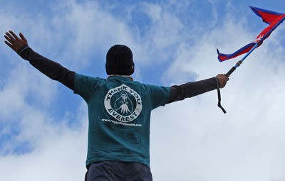 Celebrating the Everest Base Camp trek - A trekker wearing a Rugged Trails Nepal shirt holding a Nepali flag with arms wide open against a blue sky.