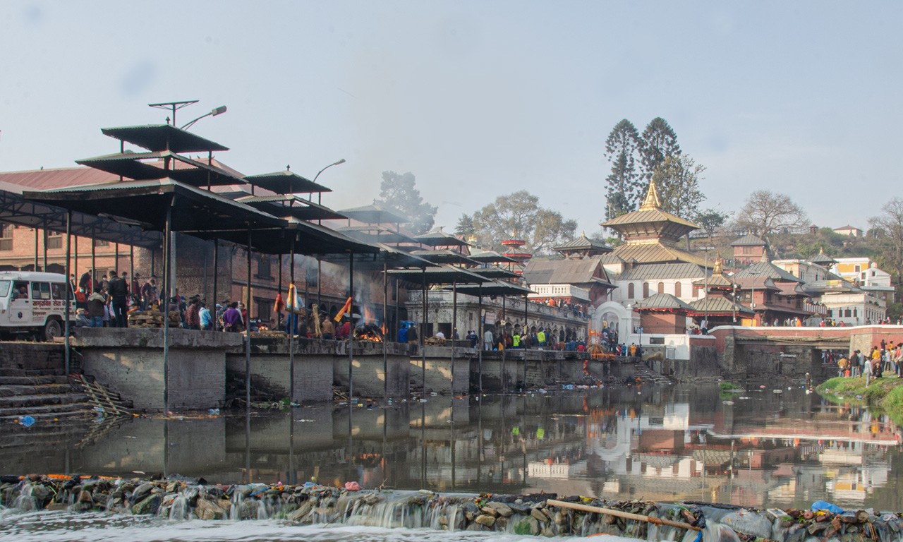 A wide view of the sacred cremation ghats at Pashupatinath Temple in Kathmandu, showing funeral pyres along the Bagmati River with temple structures in the background.