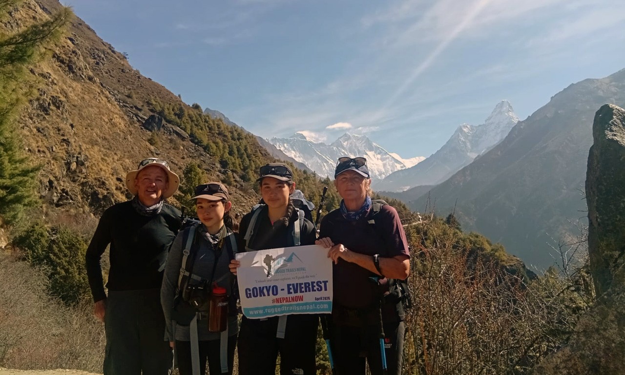Four trekkers—two men and two women—stand on a rugged mountain path in Nepal, holding a banner that reads "GOKYO - EVEREST #NEPALNOW." They are dressed in hiking gear with backpacks against a backdrop of the arid Khumbu valley and the towering, snow-capped peaks of the Himalayas under a clear blue sky.