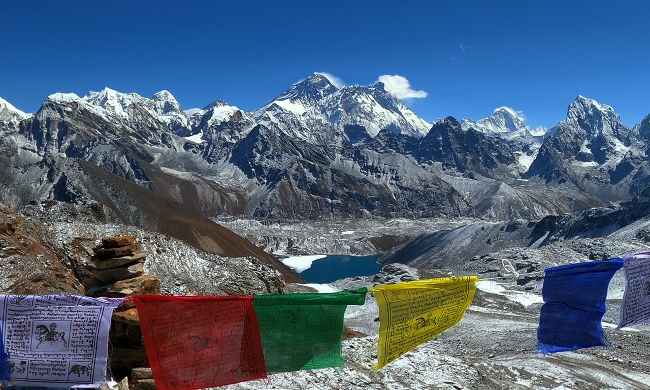 Panoramic view of Mount Everest, Lhotse, and Makalu from Gokyo Ri viewpoint with turquoise Gokyo Lake and prayer flags in the foreground.
