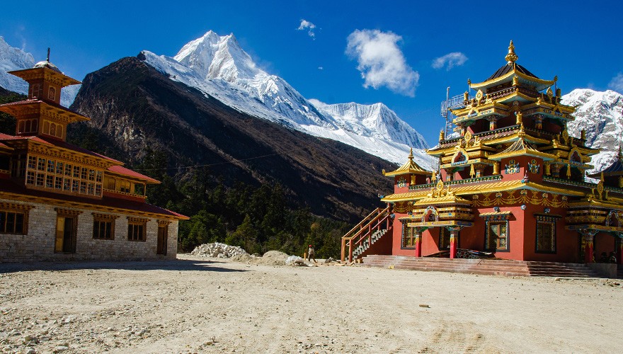The vibrant Lho Monastery with intricate Tibetan architecture set against the backdrop of the white peaks of the Manaslu massif under a clear blue sky.